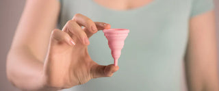 Person i ljusgrön topp som håller upp en rosa menskopp mellan fingrarna, med fokus på produkten